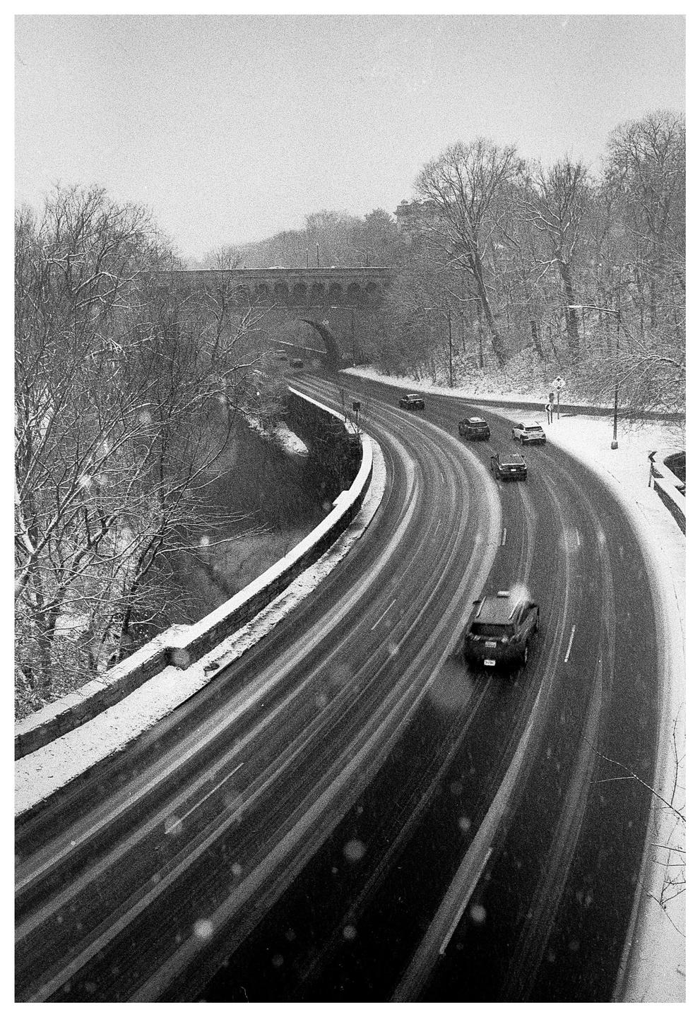 A snow-covered road with cars driving on it curves through a forest with a bridge in the background.