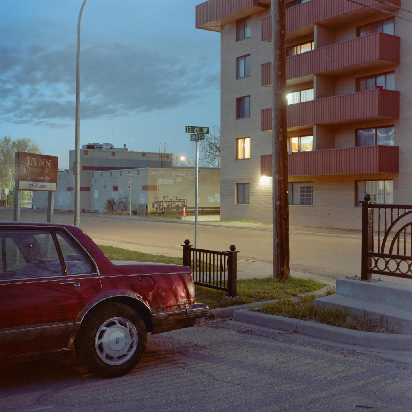 A red car is parked on a street next to an apartment building with a "no vacancy" sign.