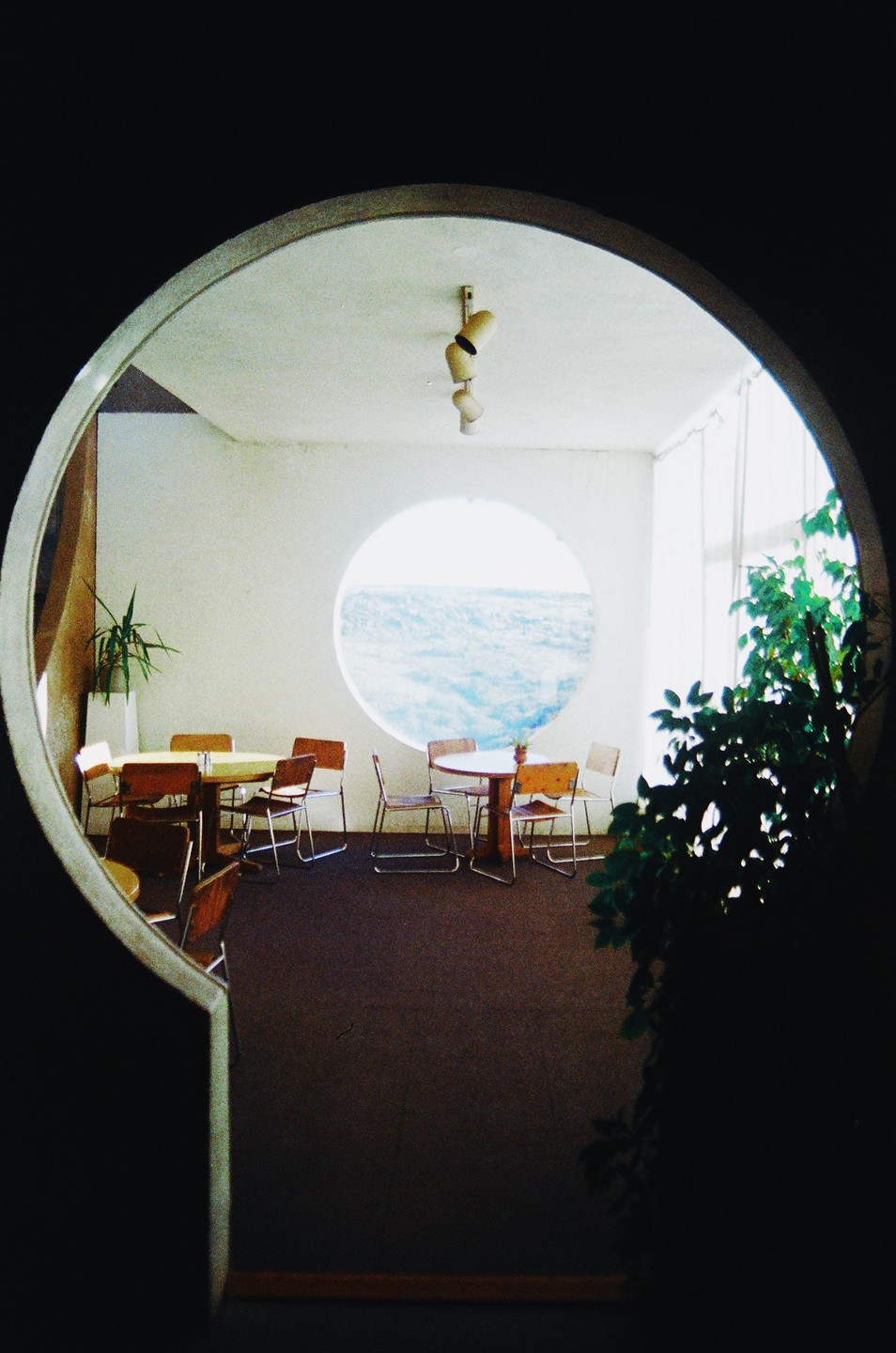 A room with tables and chairs is visible through a keyhole-shaped opening, with a circular window looking out onto the ocean.