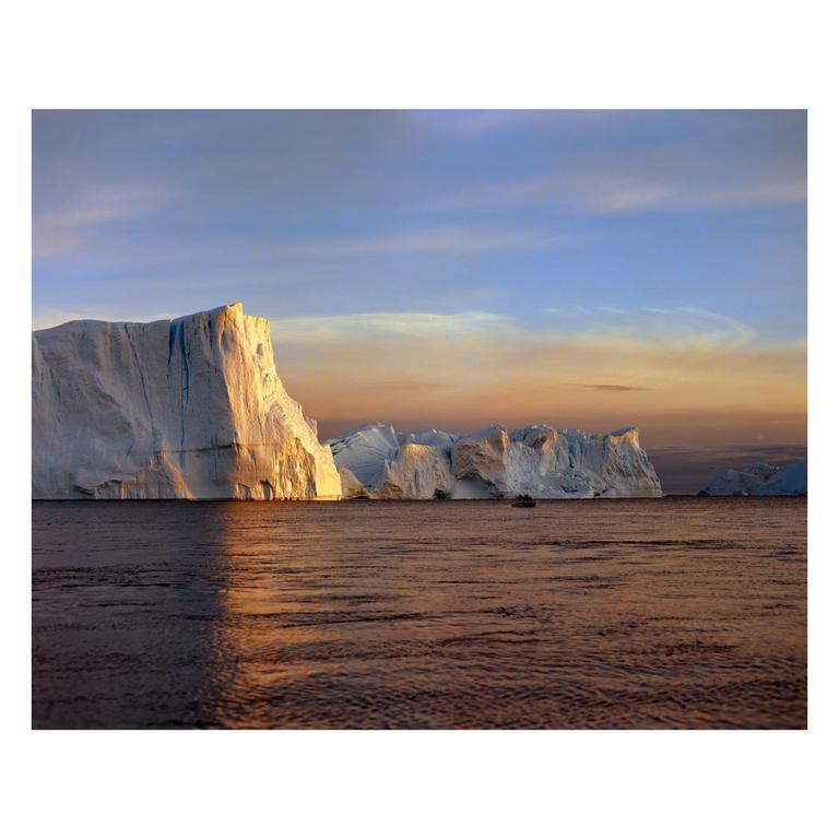 A boat floats on the water in front of a large iceberg at sunset.