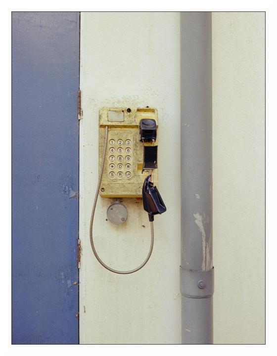 A broken yellow payphone is mounted on a white wall next to a blue door and a gray pipe.