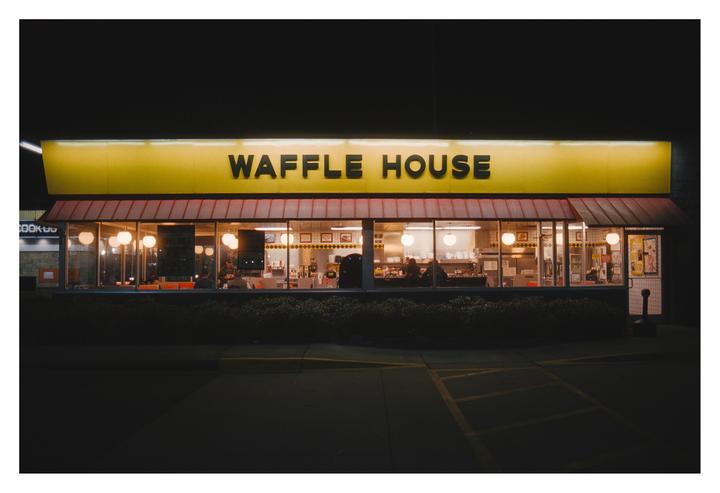 The exterior of a Waffle House restaurant is illuminated at night, with people visible inside through the windows.
