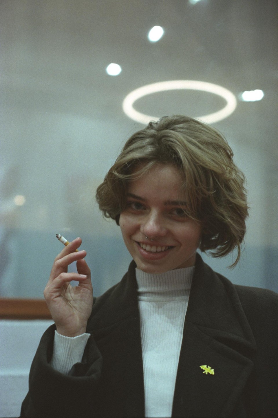 A woman with short hair and a nose piercing smiles while holding a lit cigarette.