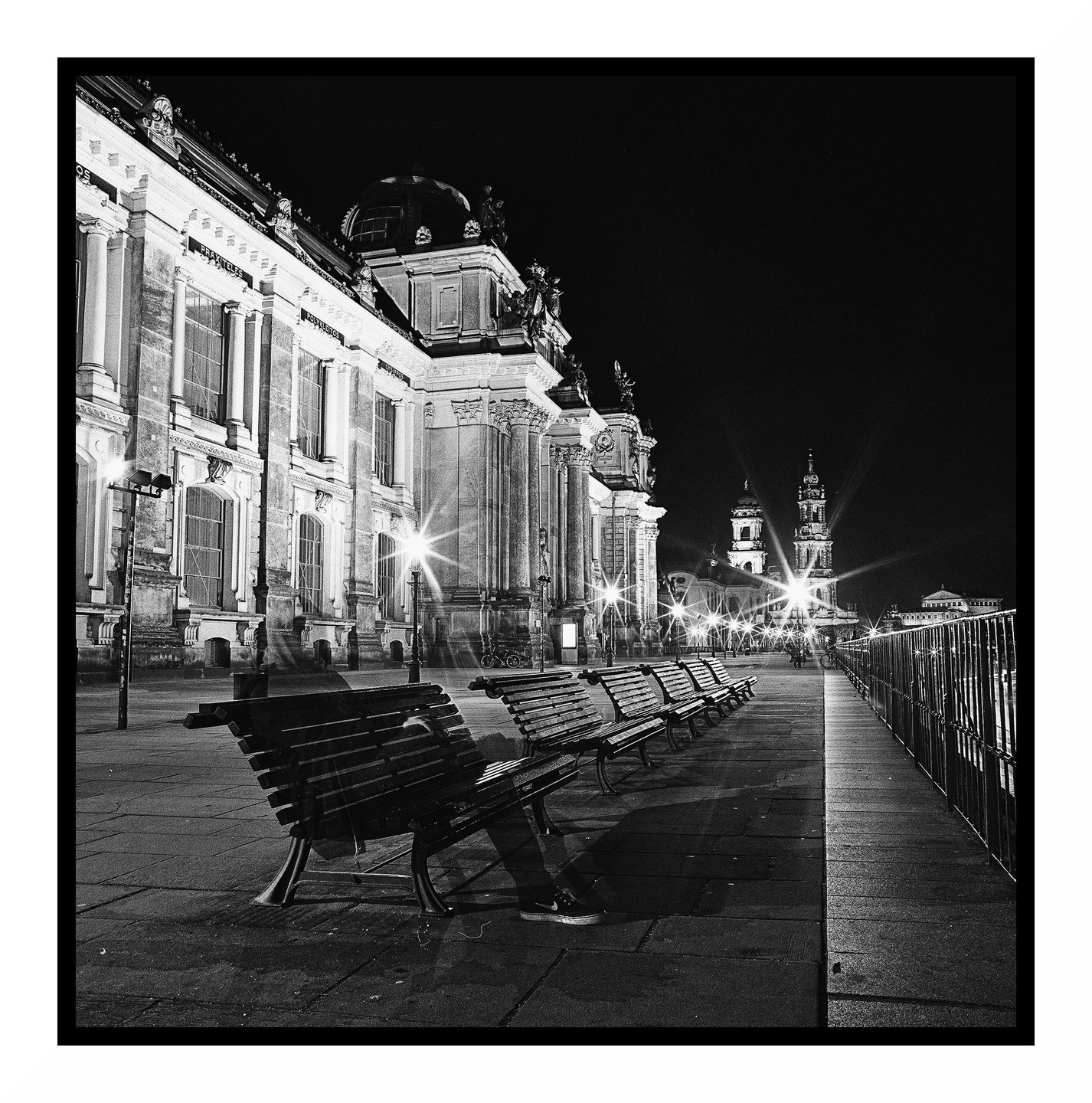 A row of empty benches sits along a promenade next to a large ornate building at night.