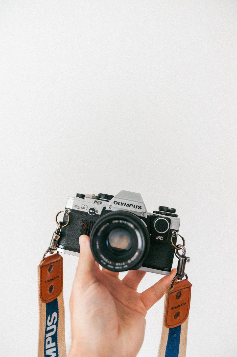 A hand holds a vintage Olympus OM10 camera with a branded strap against a white background.