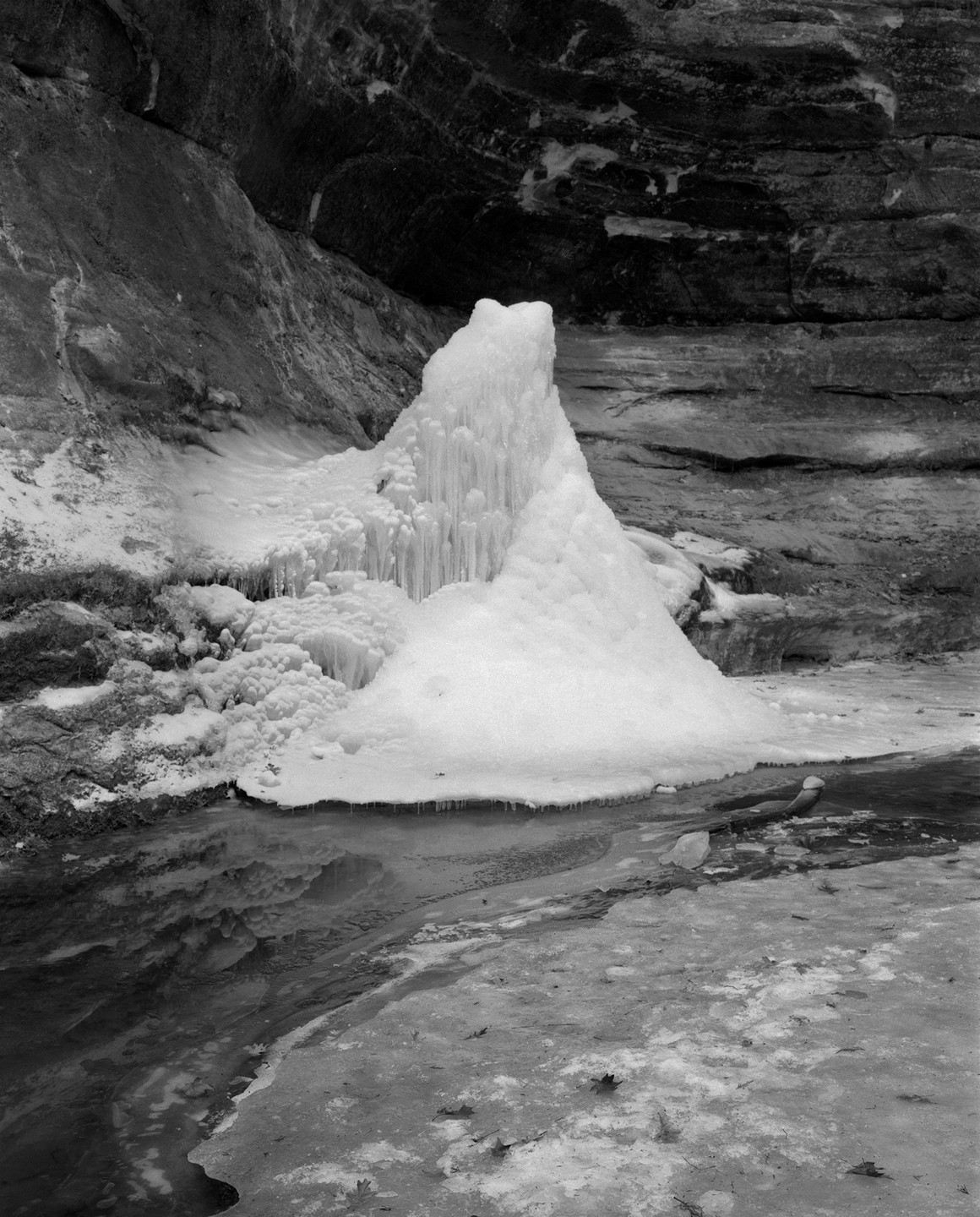 A large formation of ice and icicles stands next to a frozen stream with rock walls in the background.