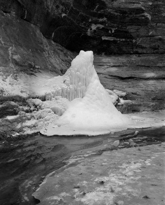 A large formation of ice and icicles stands next to a frozen stream with rock walls in the background.