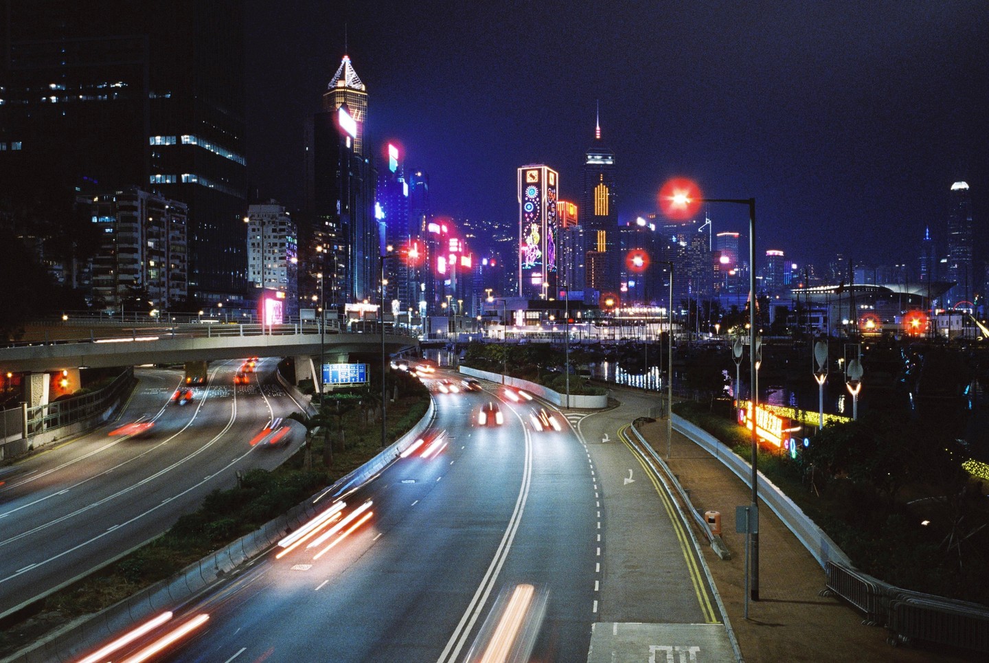 A long exposure shot captures light trails from vehicles on a multi-lane highway at night, with a city skyline illuminated by neon lights and skyscrapers in the background.