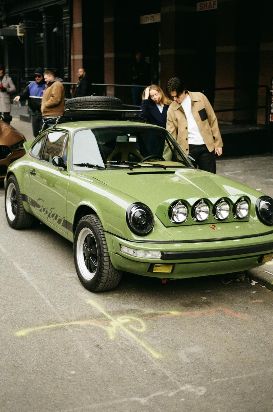 A green car with a roof rack and four auxiliary lights on the hood is parked on a street with people in the background.