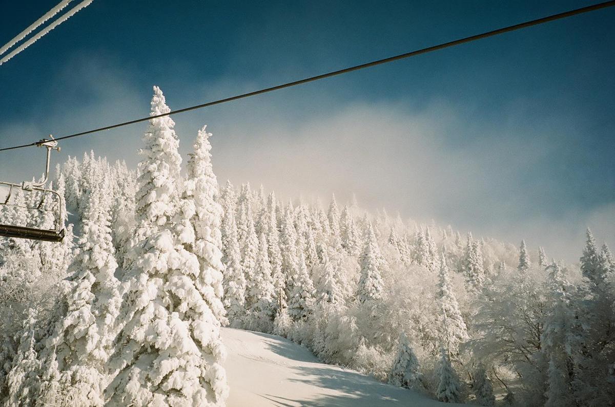 A ski lift chair is visible on the left, with snow-covered trees and a snowy slope below, under a blue sky with some clouds.