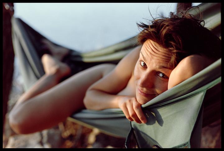 A woman with messy hair lies in a hammock, looking directly at the viewer.