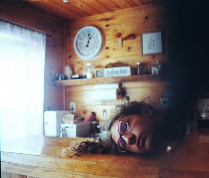 A woman with curly hair and glasses is lying on a wooden counter in a room with wood paneling.