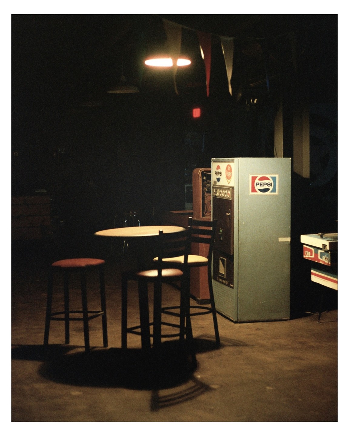 A table with two stools and a chair stands next to a Pepsi machine in a dimly lit room.