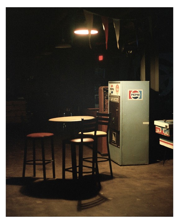 A table with two stools and a chair stands next to a Pepsi machine in a dimly lit room.