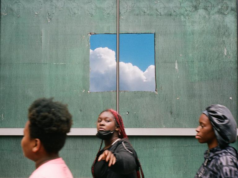 Three people walk past a green wall with a square hole revealing a blue sky with clouds.