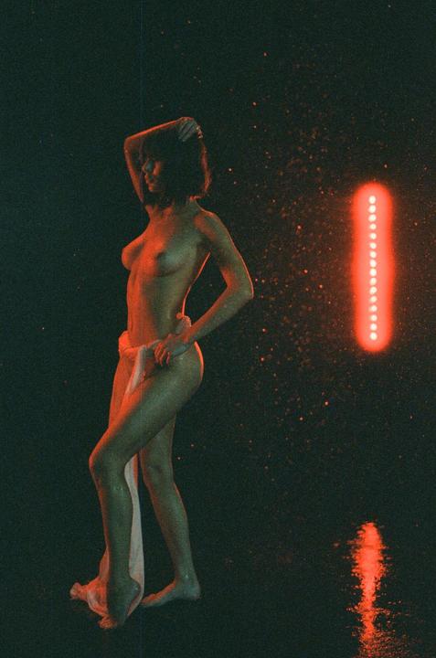 A woman stands partially nude with a white cloth draped around her hips, illuminated by red light and surrounded by water droplets.