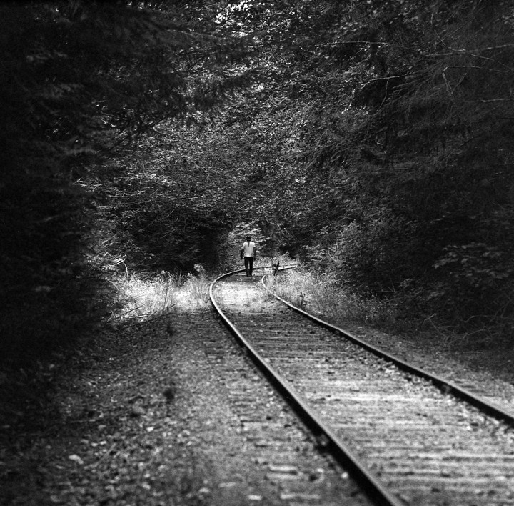 A man walks along a curving railway track through a forest.
