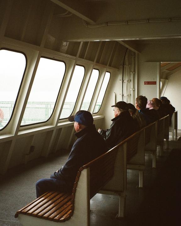 People sit on wooden benches inside a ferry, looking out of large windows.