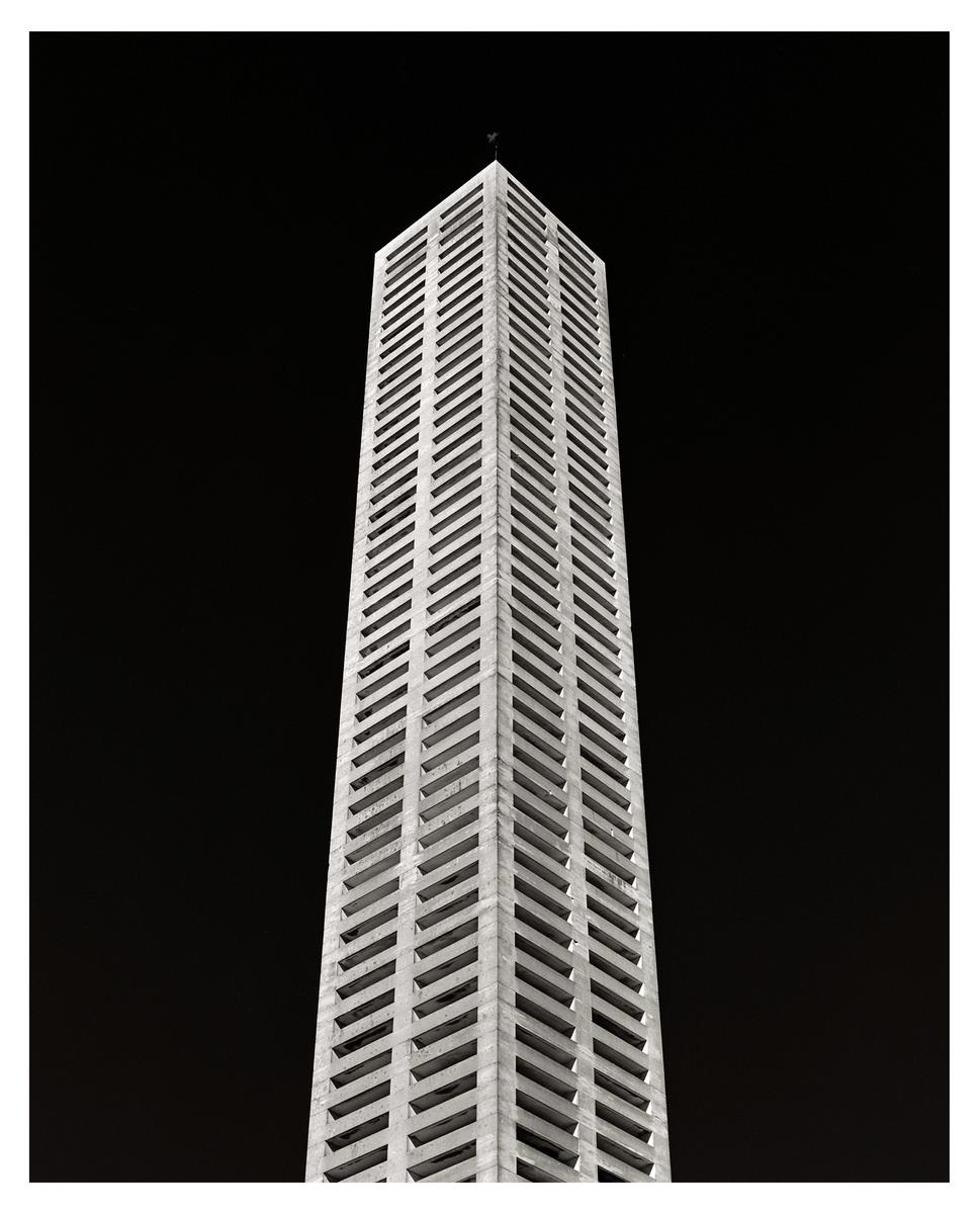 A tall, narrow building with a repeating pattern of windows rises against a dark sky.