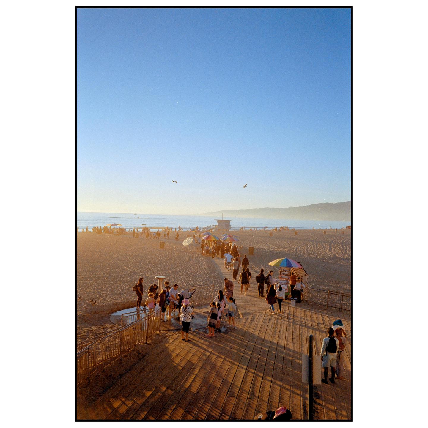 A crowd of people walk along a boardwalk and sandy beach at sunset, with mountains in the distance.