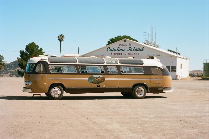A vintage brown and white bus is parked in front of a white building with "Welcome to Catalina Island Airport in the Sky" written on it.