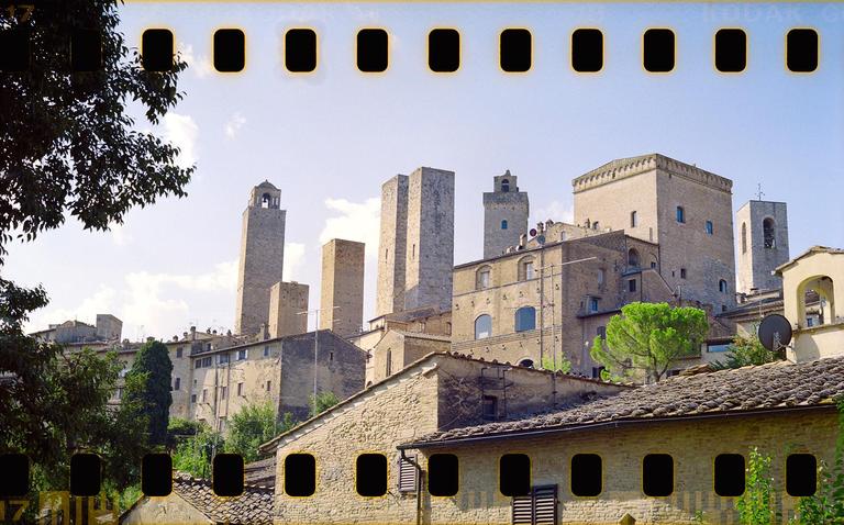 A medieval town with multiple stone towers rises above a hillside with trees and houses.