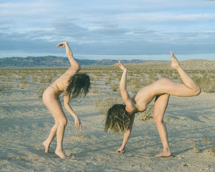 Two women pose nude in a desert landscape with mountains in the distance.