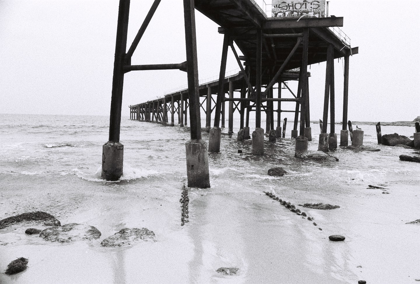 A long pier extends over the water and sand, with waves crashing around its supports.
