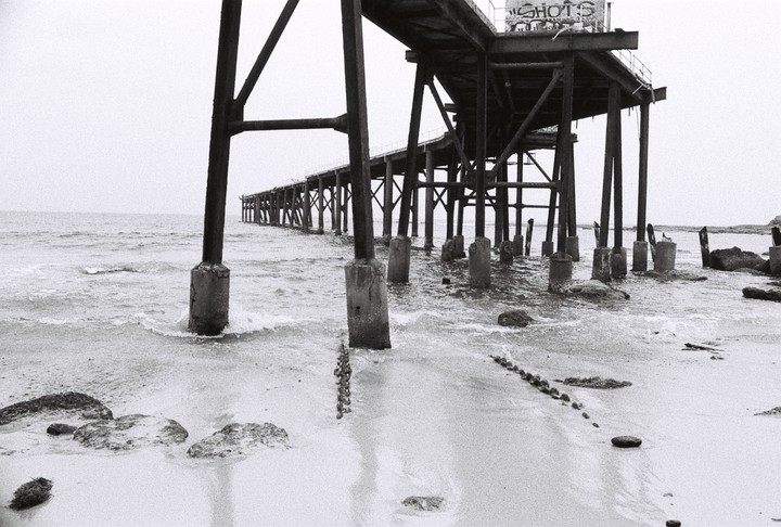 A long pier extends over the water and sand, with waves crashing around its supports.