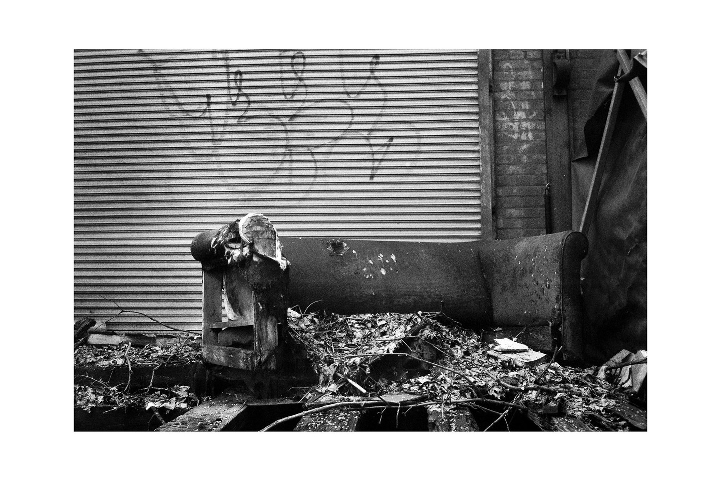 A discarded sofa sits amidst debris in front of a corrugated metal wall with graffiti.