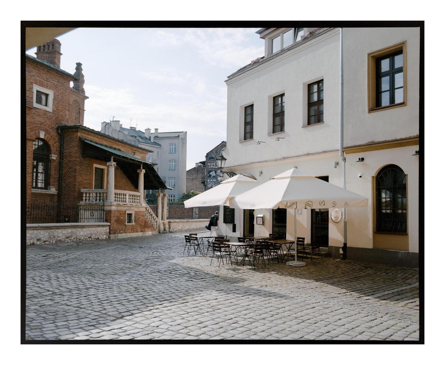 A cobblestone street is lined with buildings and an outdoor cafe with white umbrellas.