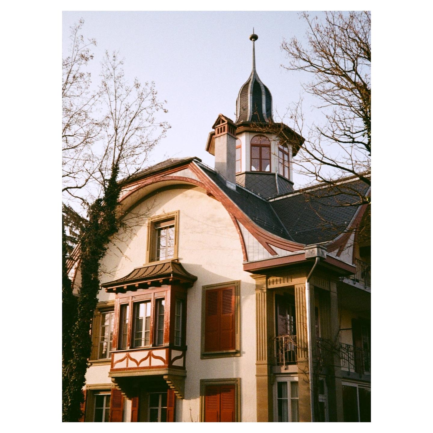 A large, ornate building with a turret and bay window stands against a clear sky, with bare tree branches framing the scene.