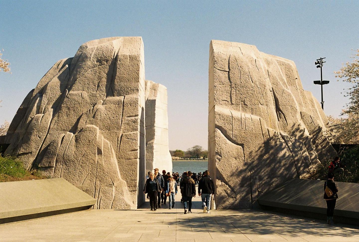 A crowd of people walk through a large stone monument with a body of water visible in the background.