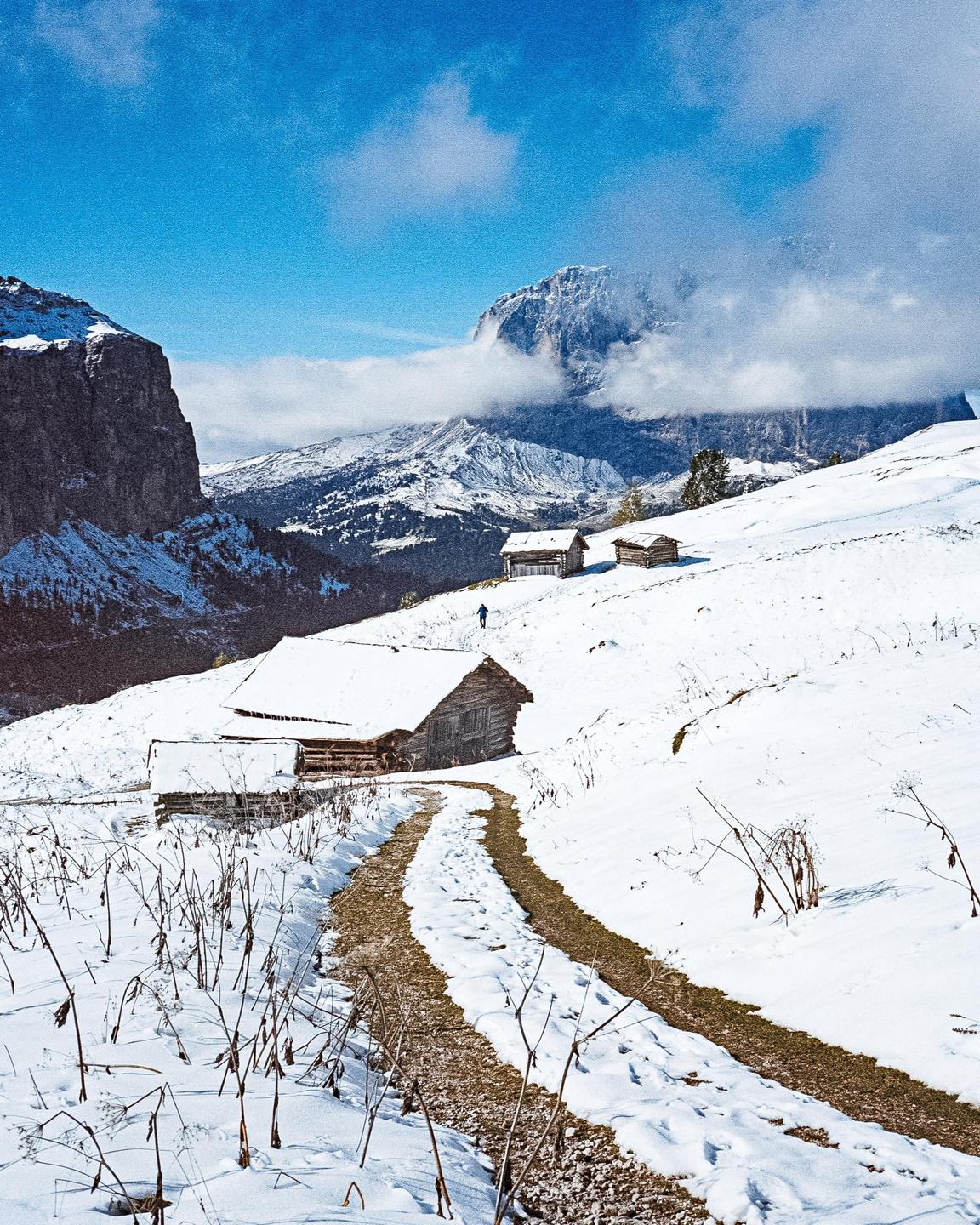 A person walks on a snowy path leading to a wooden cabin in the Italian Alps.