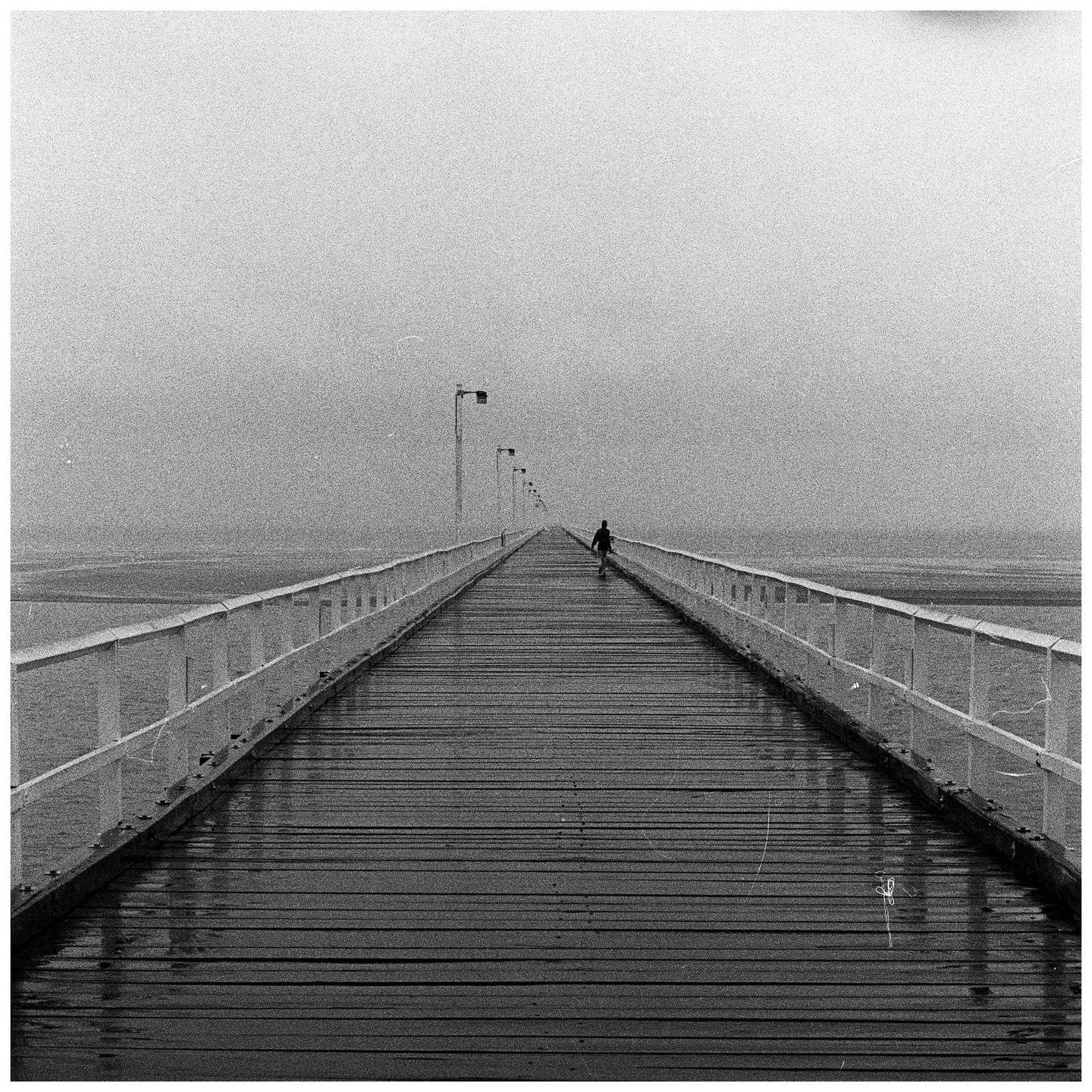 A person walks along a long pier extending into the distance under a hazy sky.