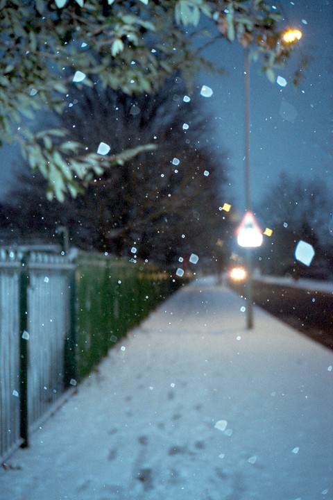 Snow falls on a sidewalk next to a fence and a street light at night.