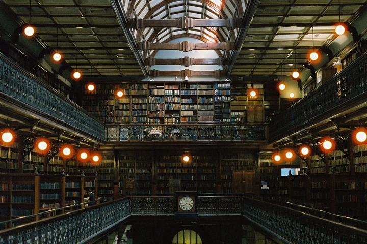 A grand library interior features multiple levels of bookshelves, ornate railings, and glowing spherical lights, with a large clock visible on the lower level.