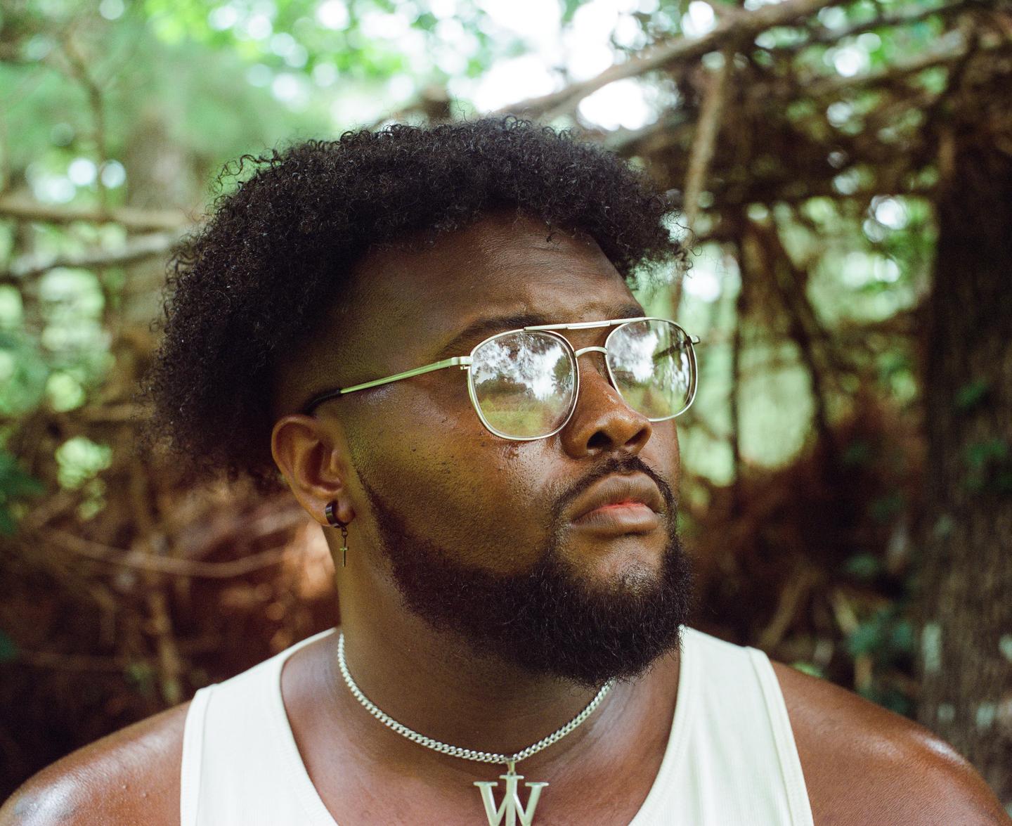 A man with a beard and sunglasses looks up while standing in a forest.