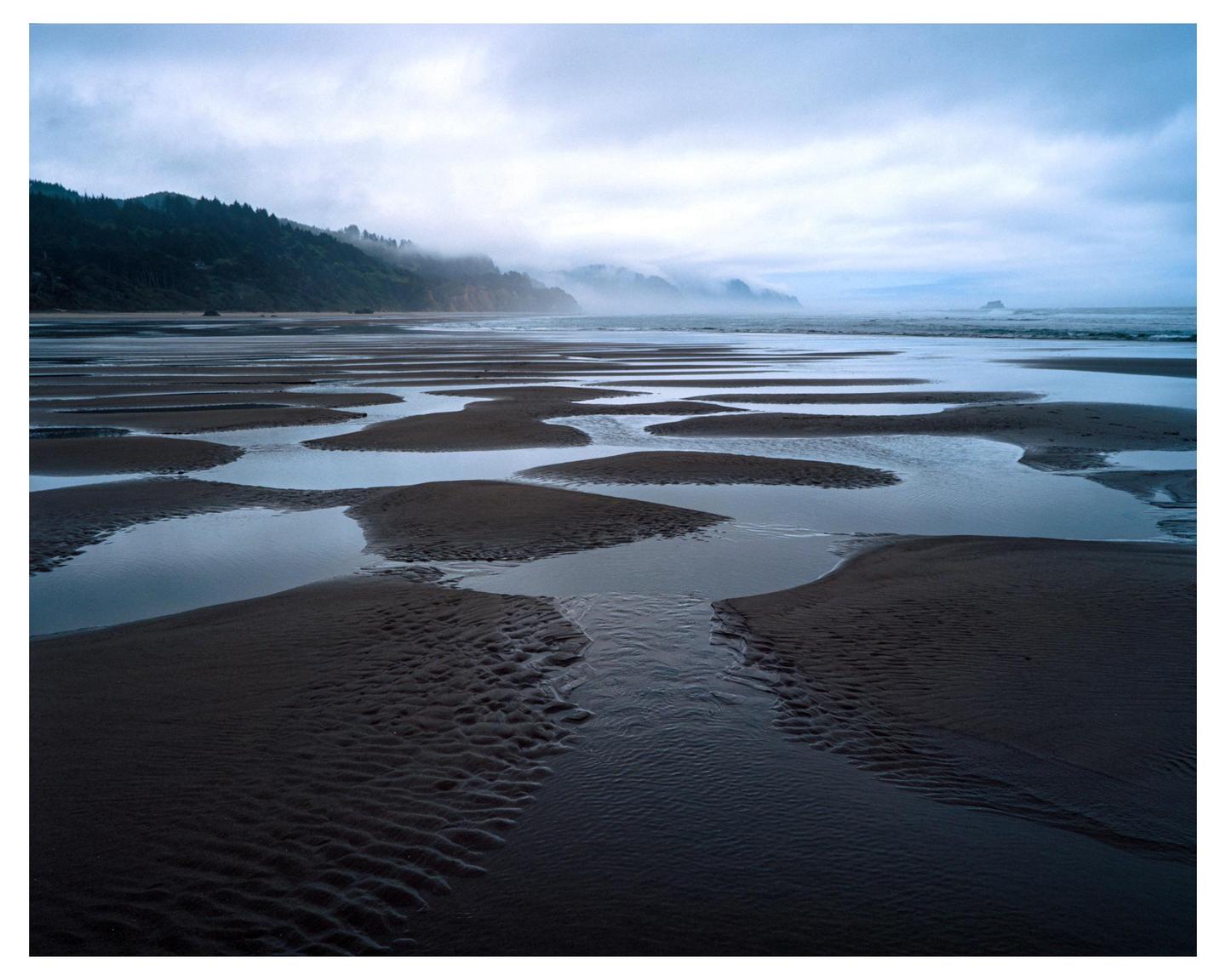 A wide expanse of beach with tidal pools and sand patterns stretches towards a misty coastline under a cloudy sky.