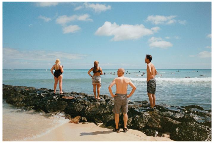 Four people stand on a rocky outcropping at the edge of a beach, looking out at the ocean where many people are swimming and surfing.