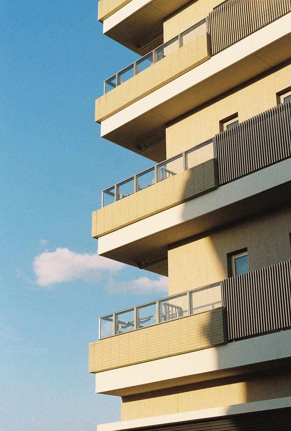 The corner of a modern building with balconies is shown against a clear blue sky with a single cloud.