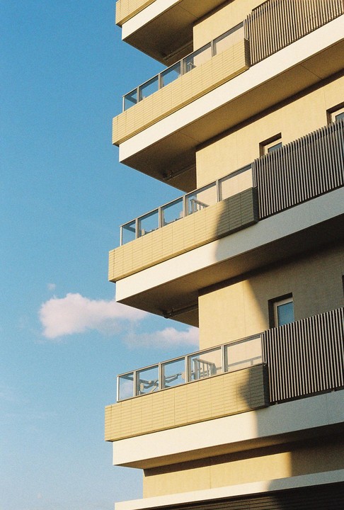 The corner of a modern building with balconies is shown against a clear blue sky with a single cloud.