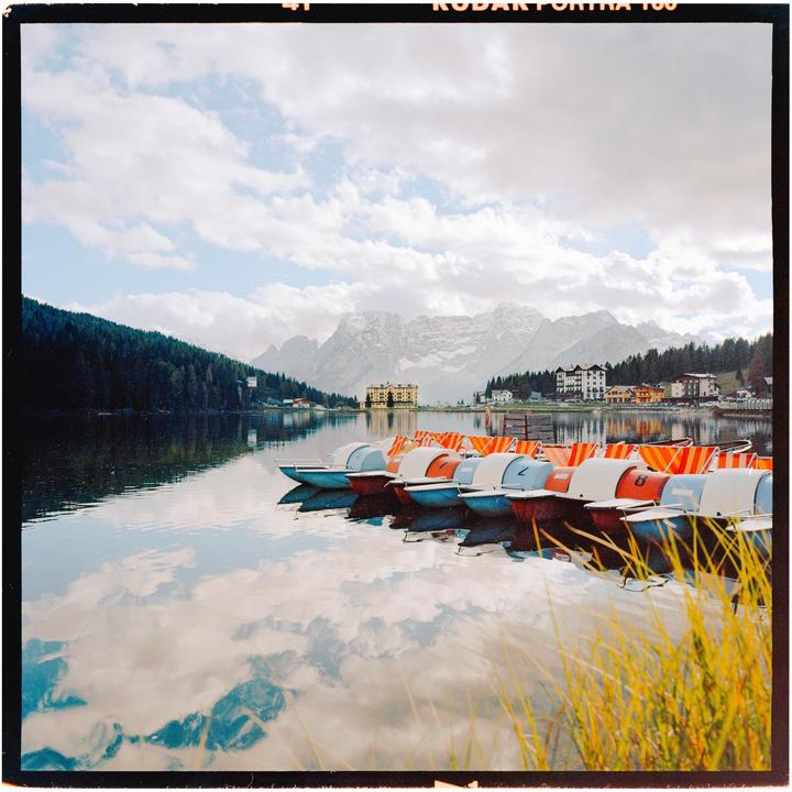 A line of colorful paddle boats floats on a calm lake with mountains and buildings in the background.