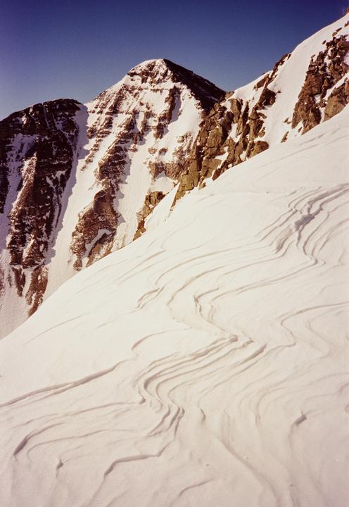 A snow-covered mountain peak rises against a clear blue sky, with textured snow in the foreground.