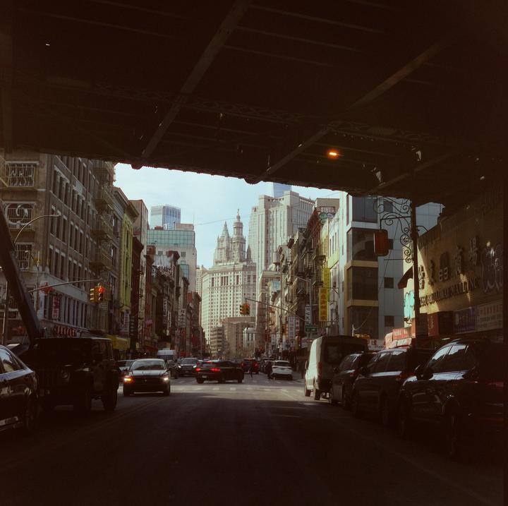 A street in a city is viewed from underneath an overpass, with cars and buildings lining the road.
