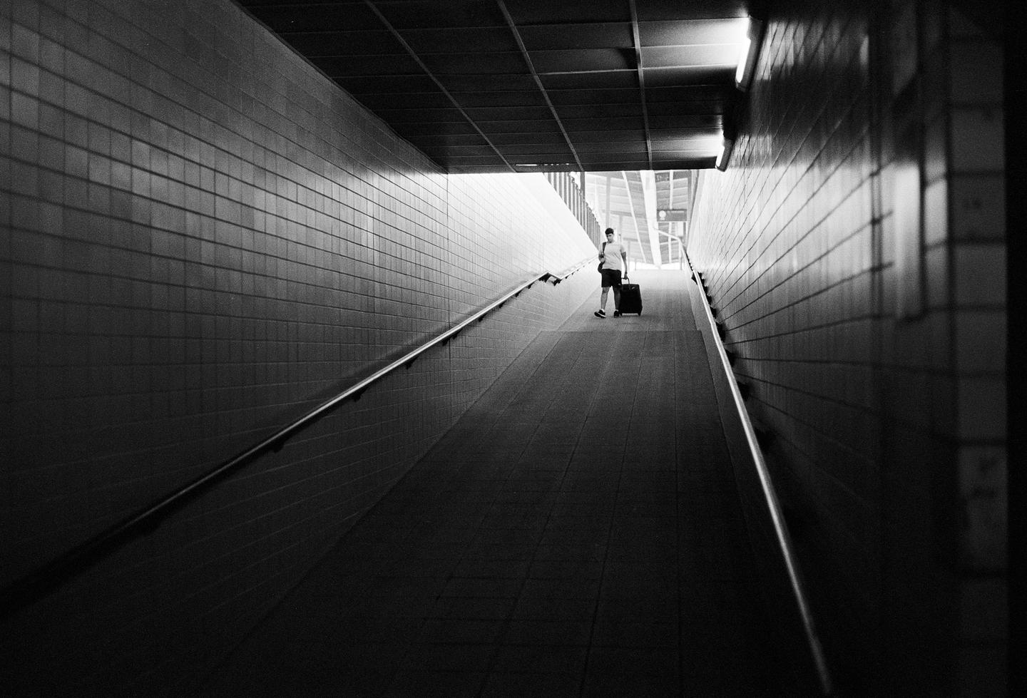 A man pulls a suitcase up a long, dark, tiled ramp toward a bright exit.