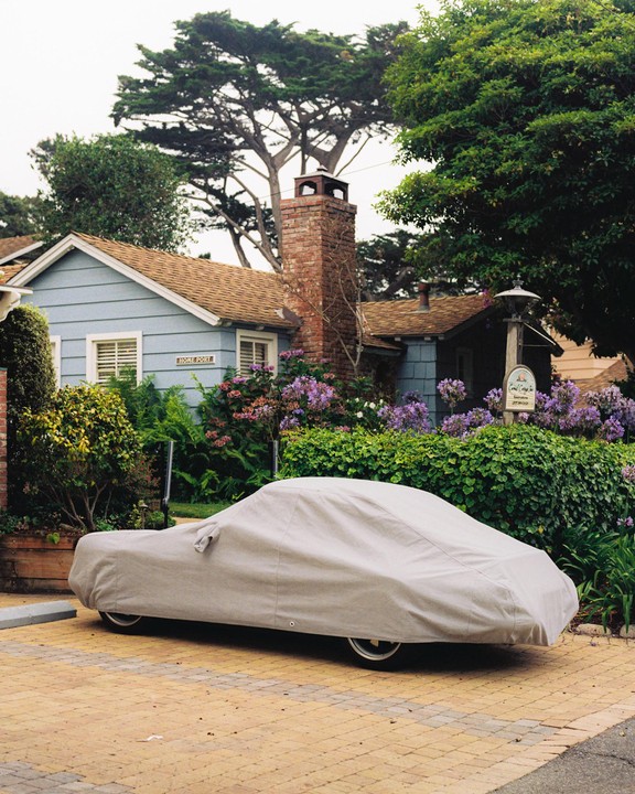 A car covered with a light-colored tarp is parked on a brick driveway in front of a house with a brick chimney and purple flowers.