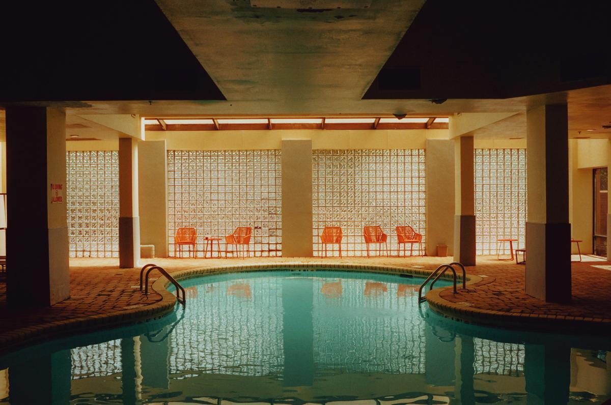An indoor swimming pool with orange chairs and tables is surrounded by glass block walls.