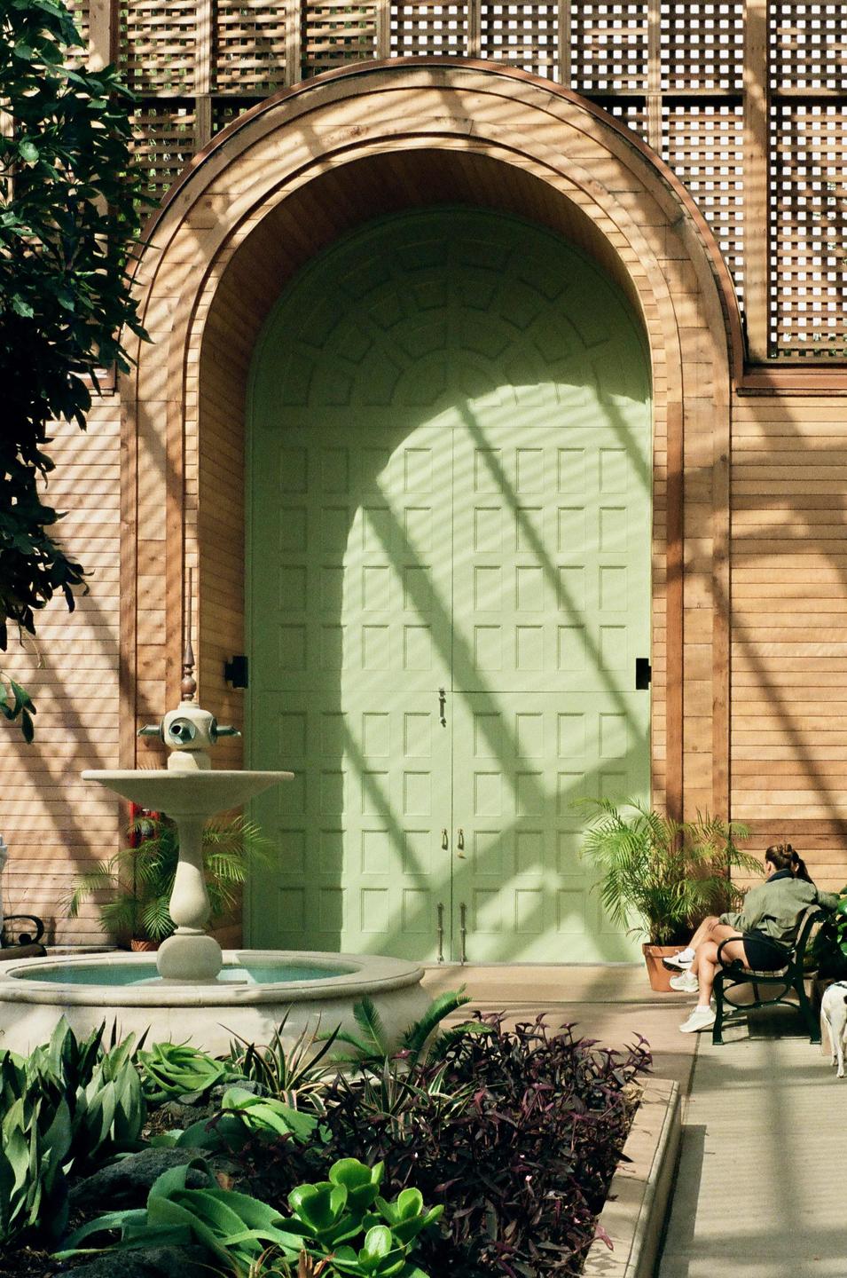 A woman sits on a bench next to a fountain and a large green door with shadows cast across it.