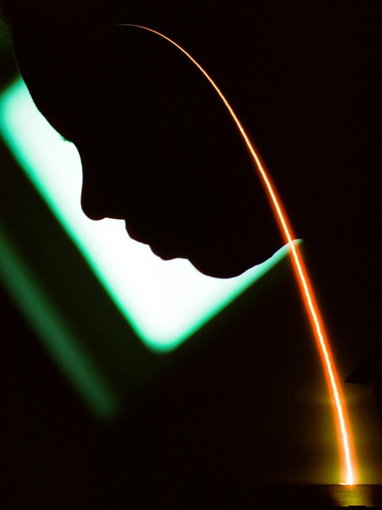 A silhouette of a person's head is visible against a bright green light, with a long orange light trail curving from the top of the frame to the bottom right.